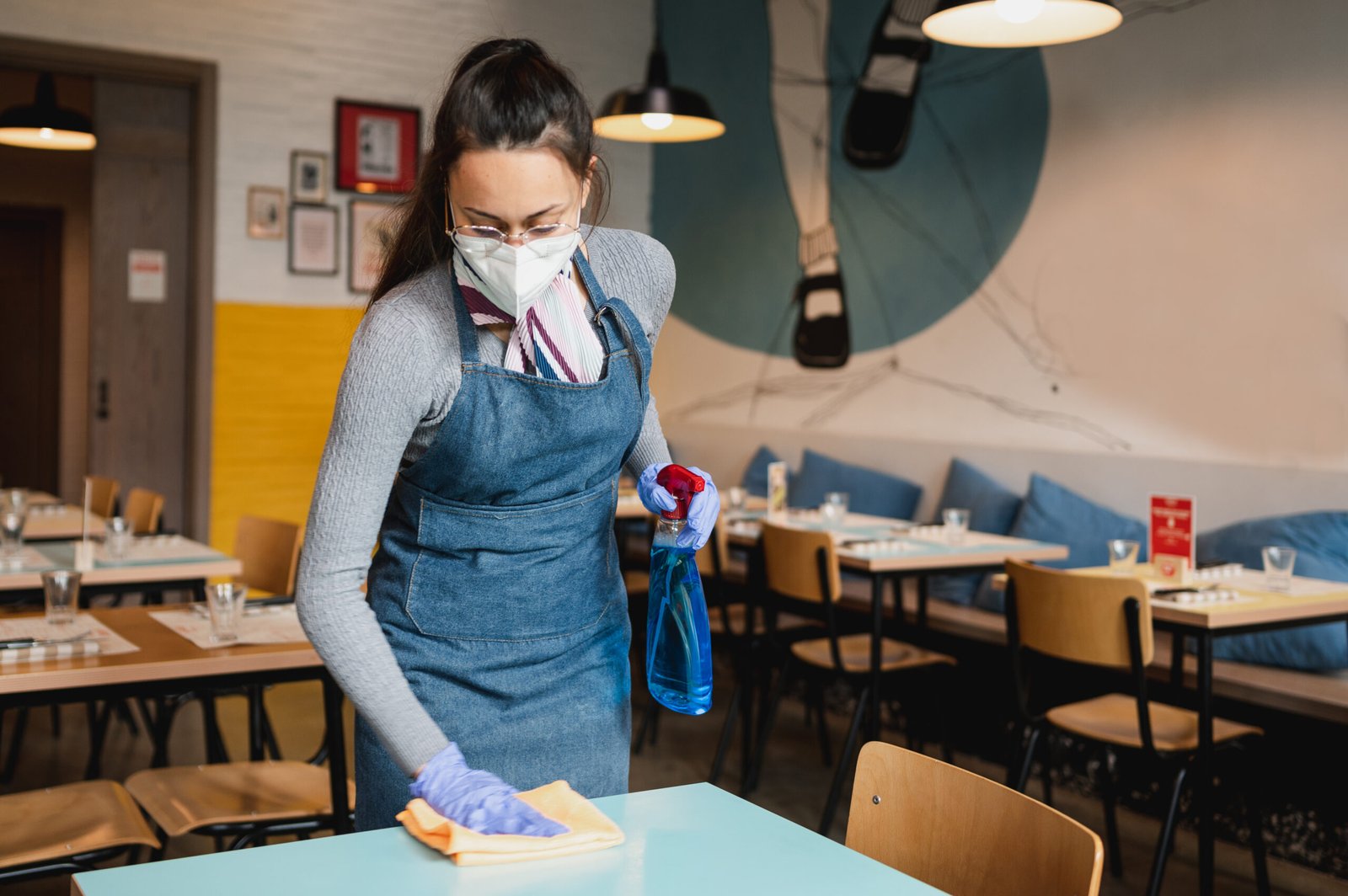 young waitress with apron, face mask and gloves cleaning tables